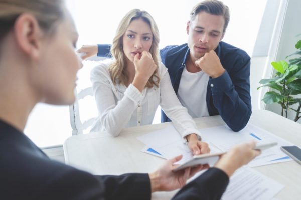 Real estate agent with couple looking through documents. The agent is holding a digital tablet showing it to the clients. Couple are casually dressed. They are looking concerned and worried and upset. Over the shoulder view.